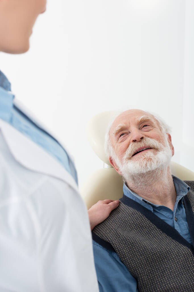 усміхнений старший пацієнт лежить у стоматологічному кріслі з рукою стоматолога на плечі
 - Фото, зображення
