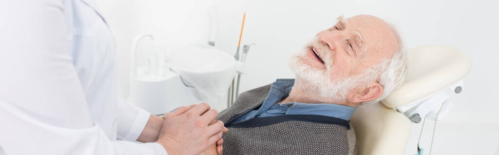 ευγνώμων ηλικιωμένος ασθενής σε οδοντιατρική καρέκλα κρατώντας τα χέρια με οδοντίατρο, πανό - Φωτογραφία, εικόνα