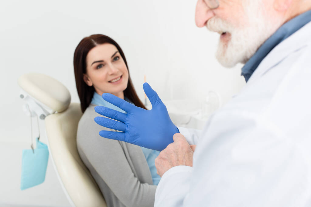 hammaslääkäri säätää lateksikäsineitä ennen hammastuolissa istuvan potilaan tutkimista - Valokuva, kuva