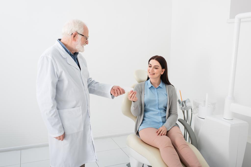dentiste senior donnant la carte vide à une femme souriante dans une chaise dentaire à la clinique  - Photo, image