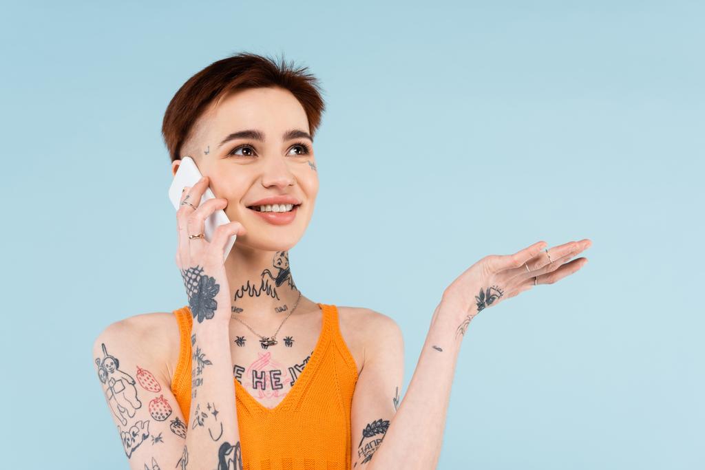 Neşeli ve dövmeli bir kadın akıllı telefonda konuşurken mavi renkte izole edilmiş bir şekilde el kol hareketi yapıyor.  - Fotoğraf, Görsel