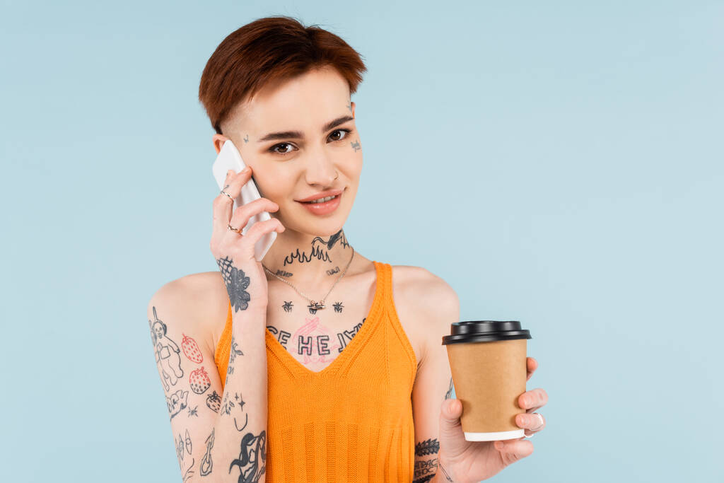 笑顔でタトゥー入りの女性がスマホで話しながらコーヒーを飲みながら青で隔離され - 写真・画像