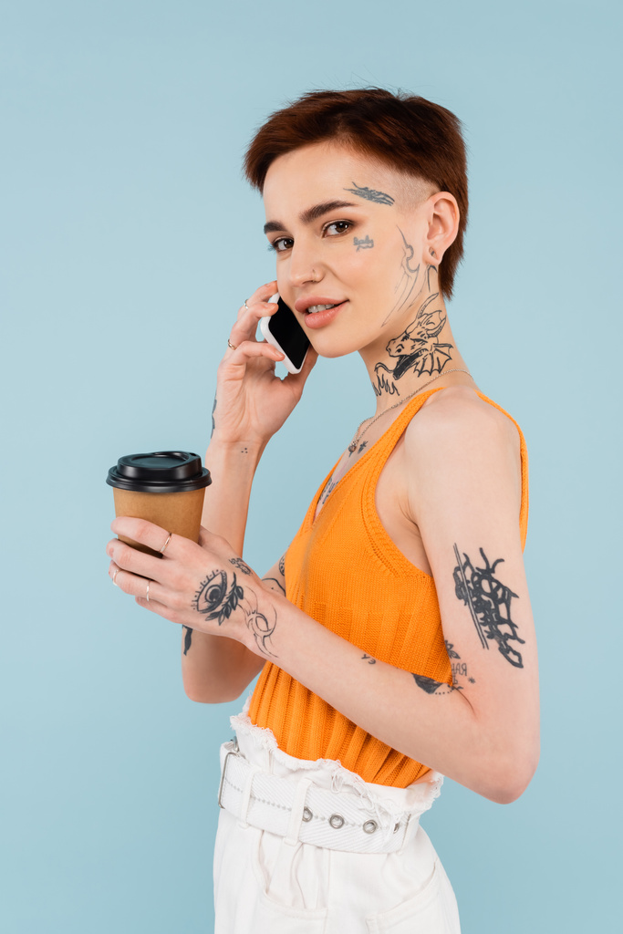 vrolijke en getatoeëerde vrouw praten op smartphone terwijl het houden van koffie te gaan geïsoleerd op blauw - Foto, afbeelding