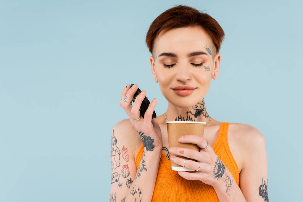Радостная и татуированная женщина держит бумажную чашку и нюхает кофе, чтобы пойти изолированы на синий - Фото, изображение