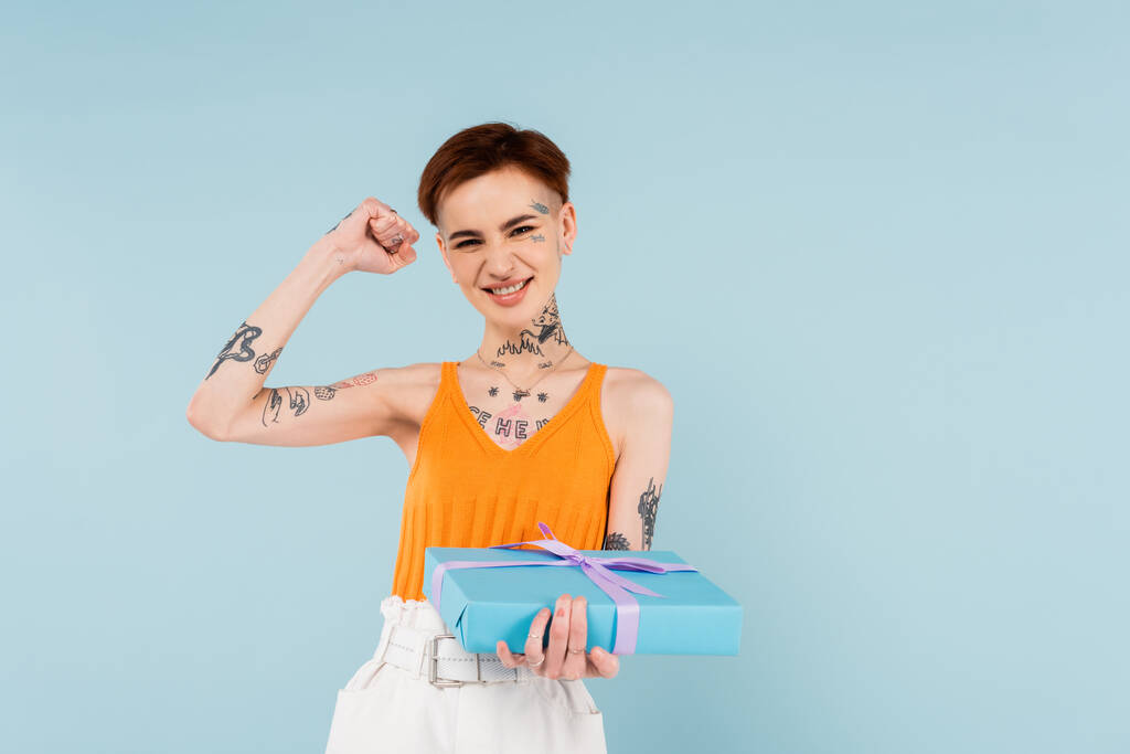 mujer excitada y tatuada sosteniendo caja de regalo envuelta aislada en azul  - Foto, Imagen