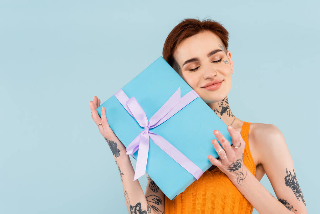 potěšená mladá a tetovaná žena drží zabalený dárek izolované na modré  - Fotografie, Obrázek