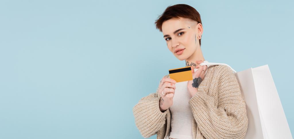 молода татуйована жінка тримає кредитну картку і сумки для покупок ізольовані на синьому, банер
 - Фото, зображення