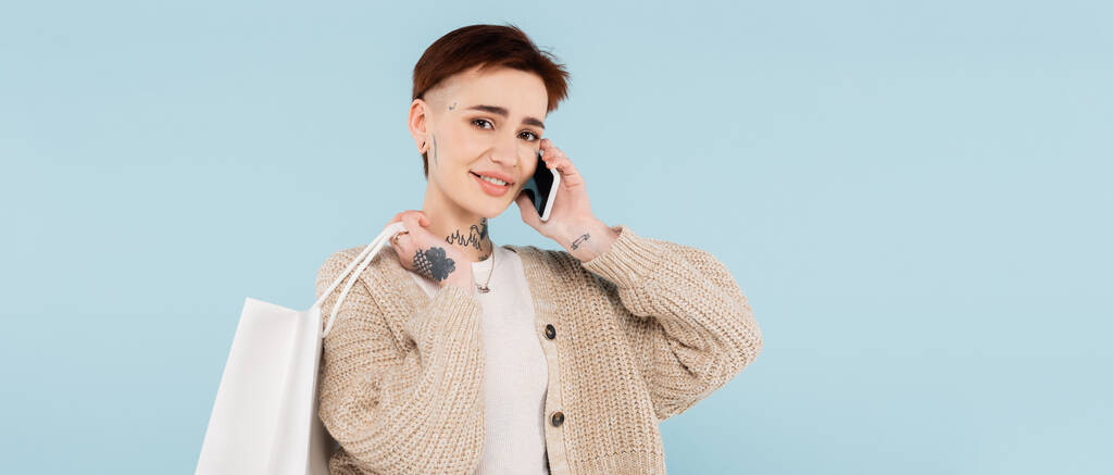 feliz joven con tatuajes sosteniendo bolsas de compras mientras habla en un teléfono inteligente aislado en azul, pancarta - Foto, imagen