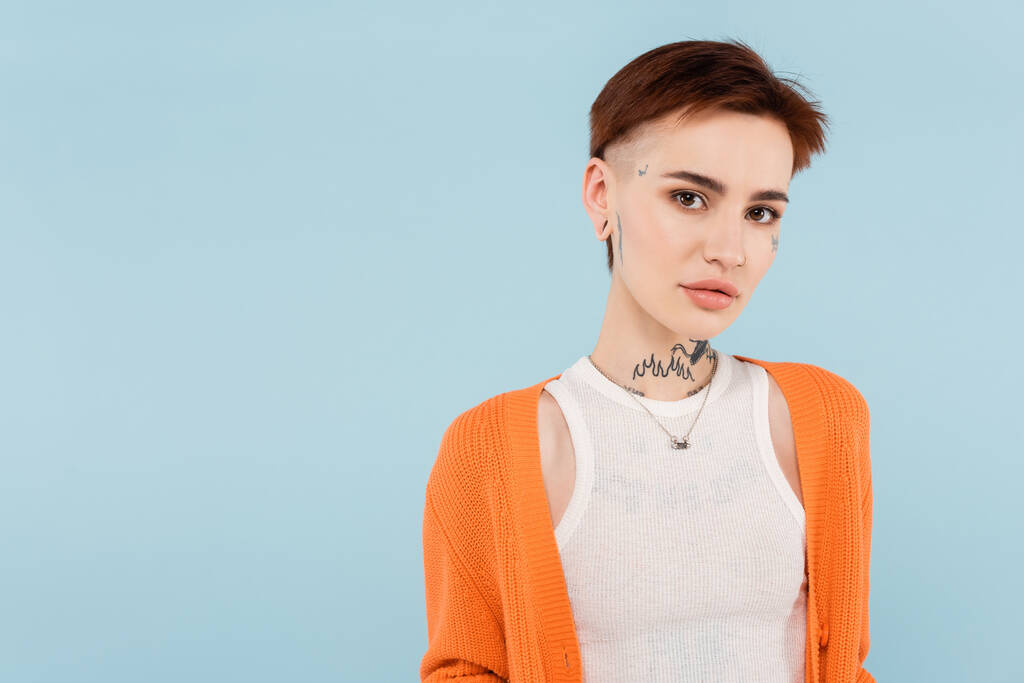 young woman with tattoos in orange cardigan looking at camera isolated on blue - Photo, Image