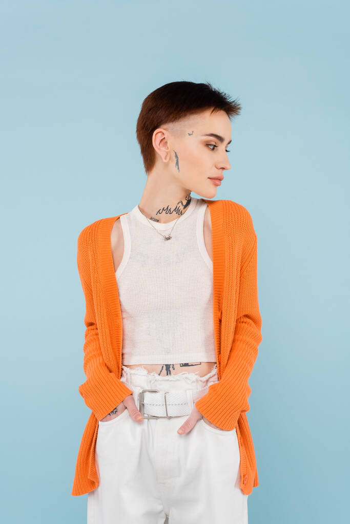  young woman with tattoos in orange cardigan posing with hands on pockets isolated on blue - Photo, Image