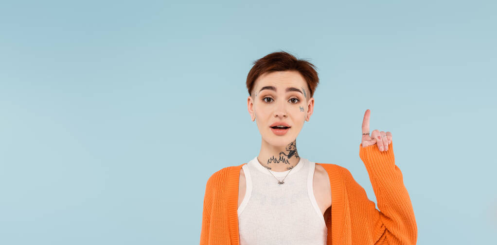stupita giovane donna tatuata in cardigan arancione rivolta verso l'alto con dito isolato su blu, striscione - Foto, immagini