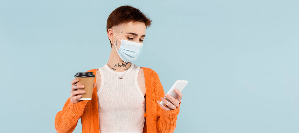 молода татуйована жінка в медичній масці за допомогою смартфона і тримає паперову чашку ізольовано на синьому, банер
 - Фото, зображення
