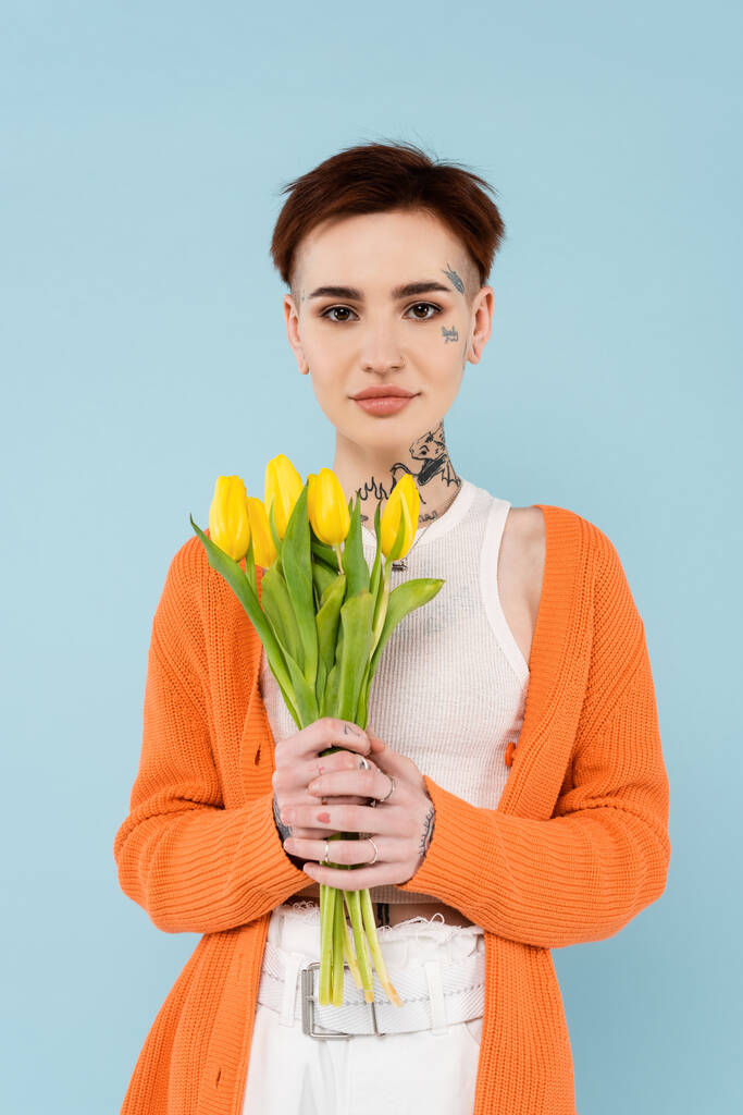 junge tätowierte Frau in orangefarbener Strickjacke mit gelben Tulpen auf blauem Grund - Foto, Bild