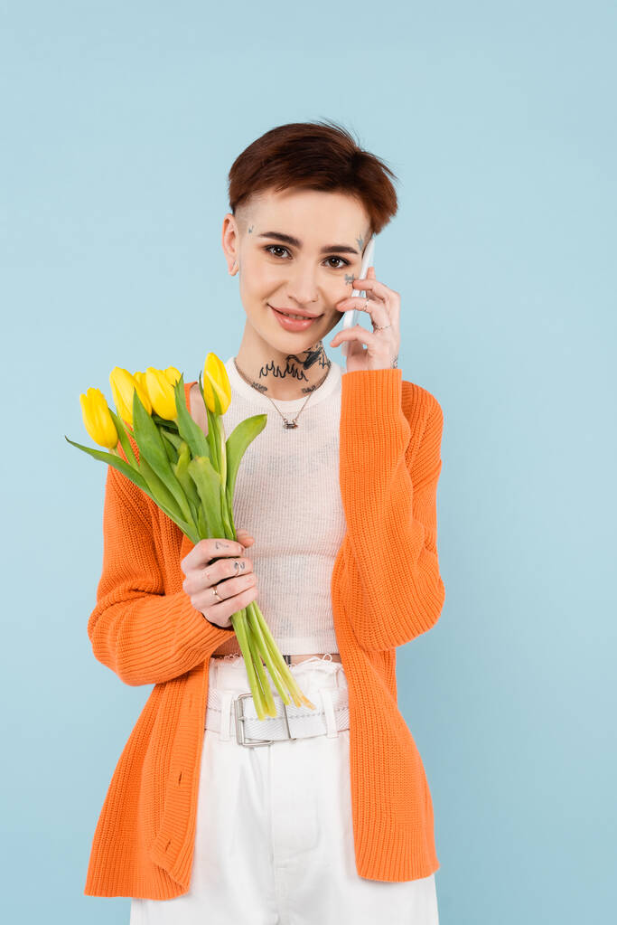 junge tätowierte Frau in orangefarbener Strickjacke mit gelben Tulpen, während sie auf dem Smartphone isoliert auf blau spricht - Foto, Bild
