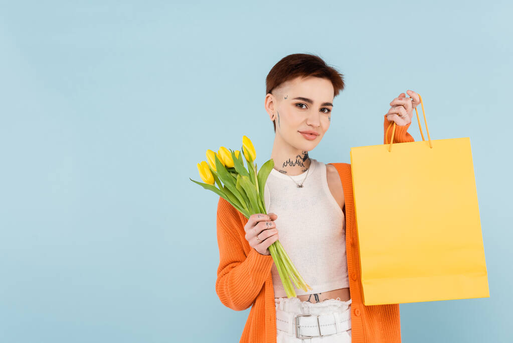 mladá tetovaná žena v oranžovém svetru drží žluté tulipány a nákupní taška izolované na modré - Fotografie, Obrázek