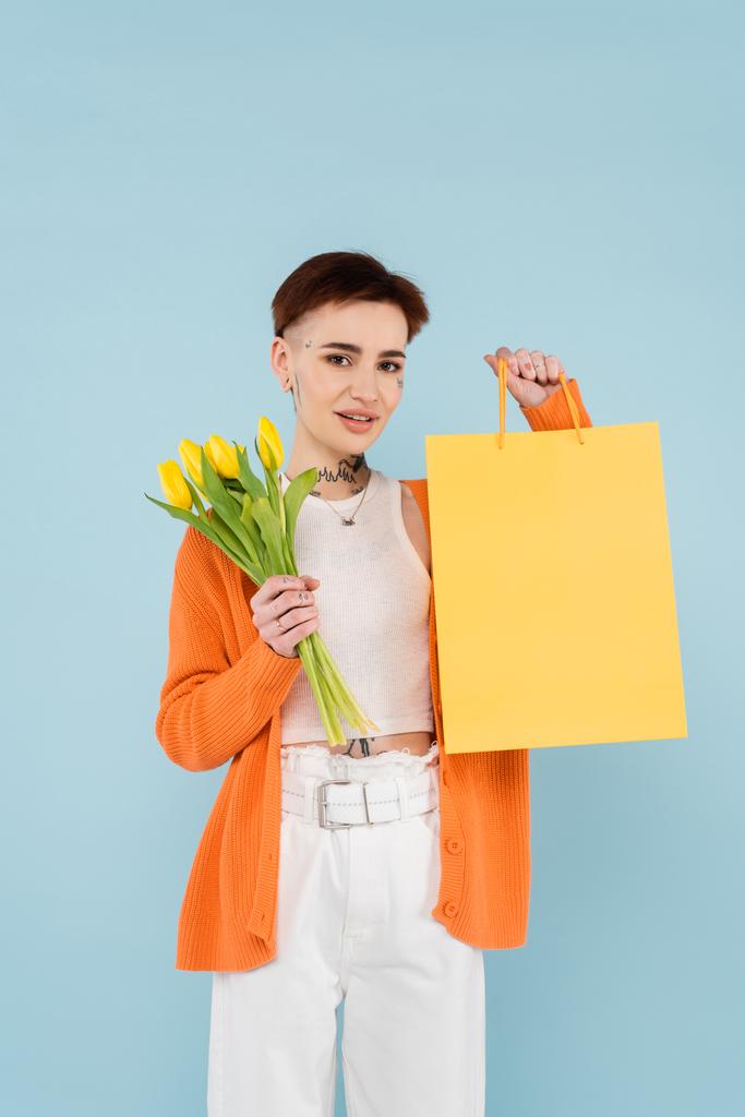 boldog nő tetovált narancs kardigán gazdaság sárga tulipán és bevásárló táska elszigetelt kék - Fotó, kép