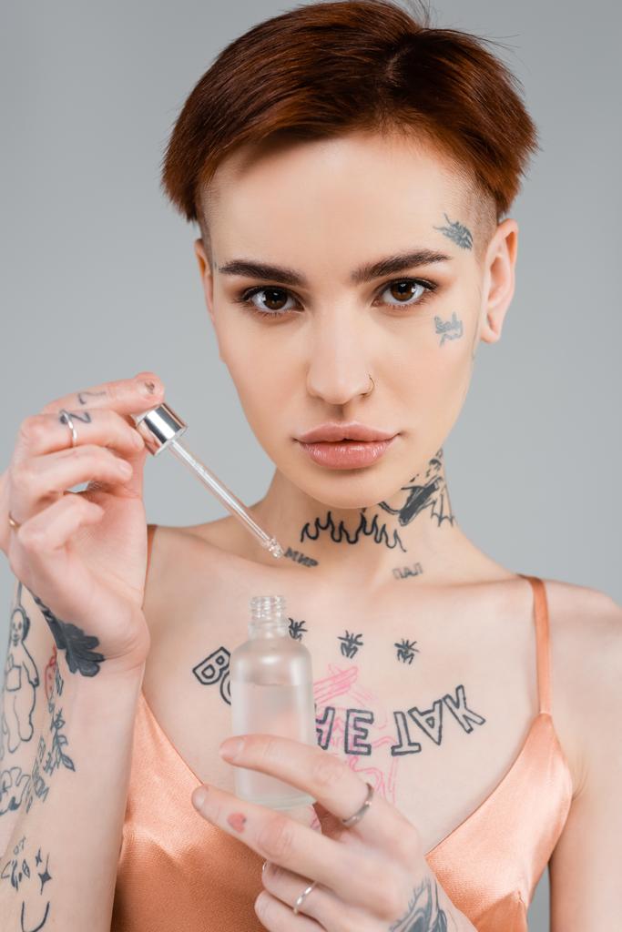 giovane donna tatuata con bottiglia di siero e pipetta isolata su grigio  - Foto, immagini