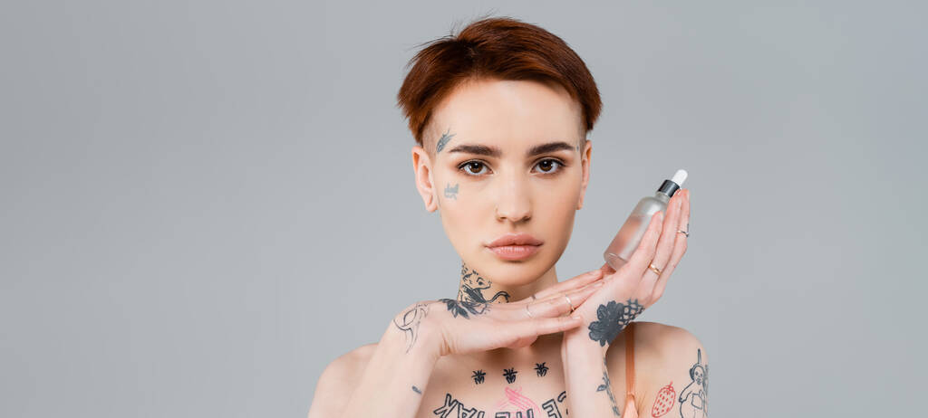 tetovaná mladá žena drží láhev se sérem izolovaným na šedé, banner - Fotografie, Obrázek
