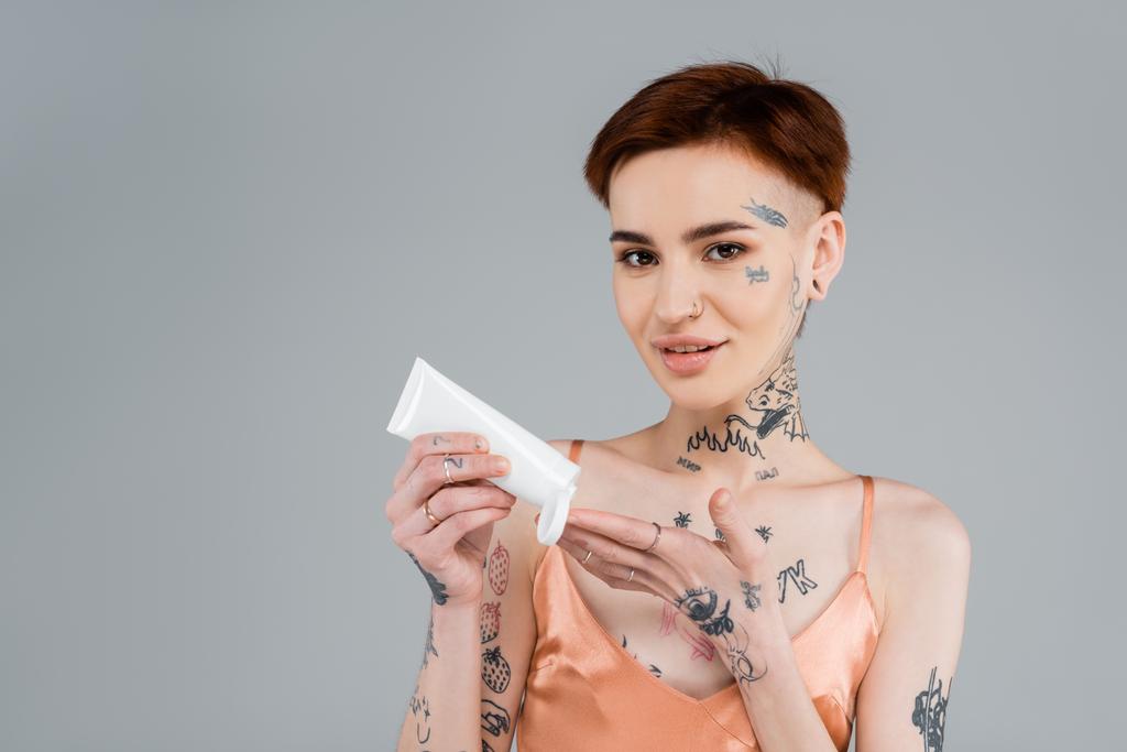 tatuoitu nuori nainen pitelee putkea ja levittää vartalovoidetta hymyillen eristettynä harmaaseen  - Valokuva, kuva