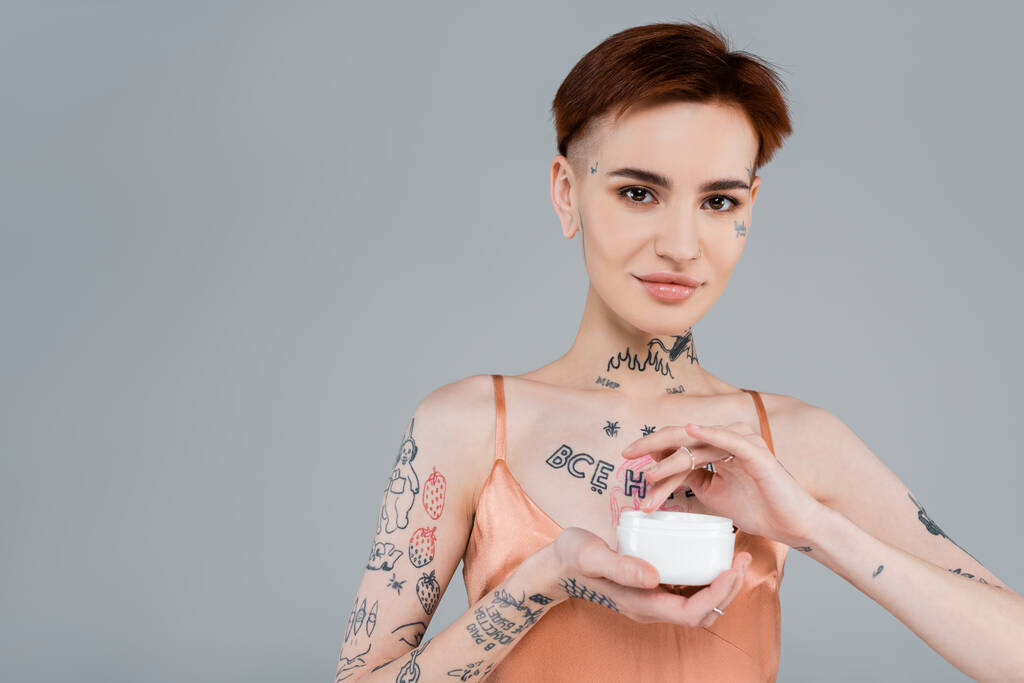 šťastná žena s tetováním a zrzavé vlasy drží bílý kontejner s kosmetickým přípravkem izolované na šedé  - Fotografie, Obrázek