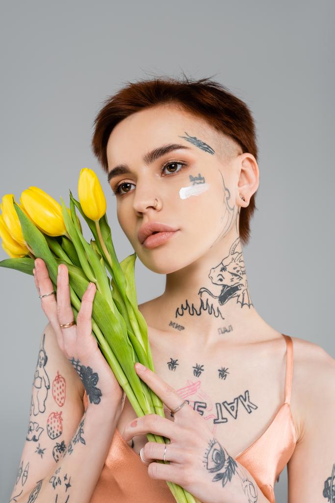 mujer joven tatuada con crema cosmética en la cara sosteniendo tulipanes aislados en gris  - Foto, imagen
