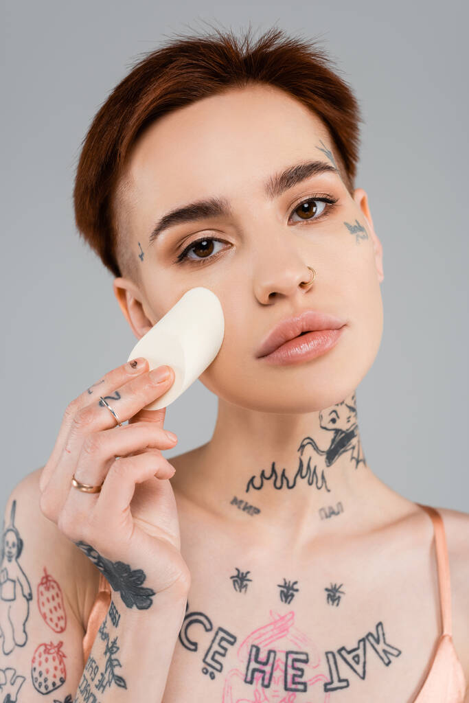 mladá tetovaná žena nanášení make-up nadace na obličej s houbou izolované na šedé - Fotografie, Obrázek