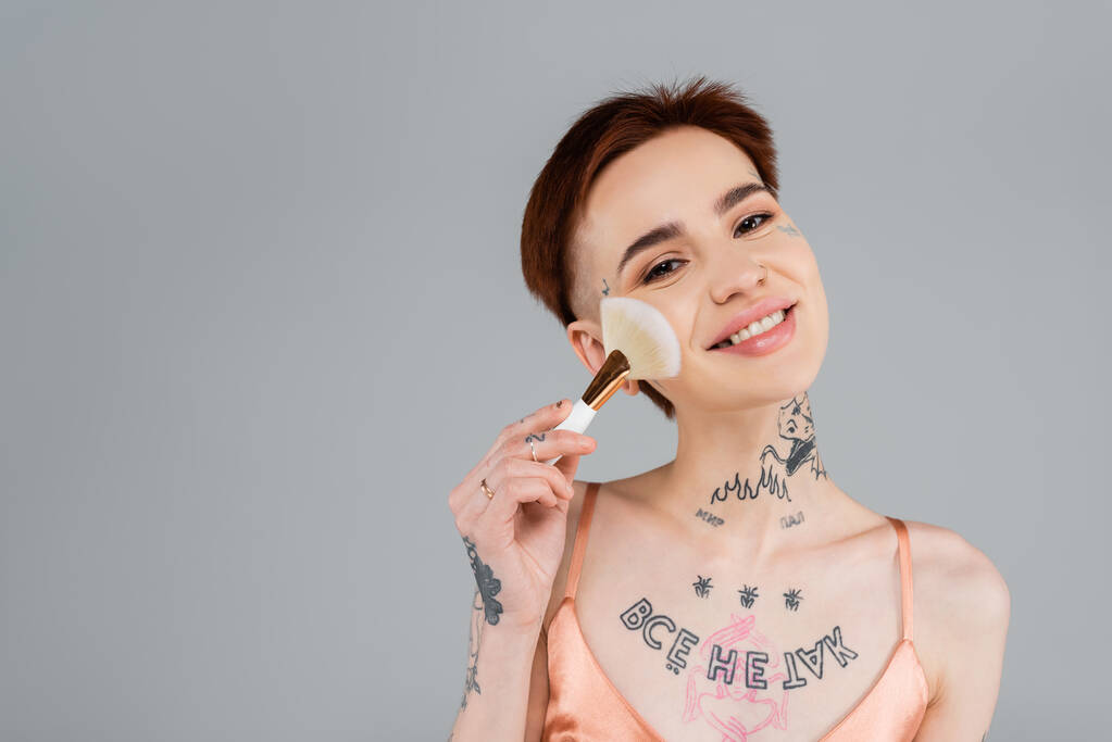улыбающаяся татуированная женщина наносит порошок для лица с косметической щеткой, изолированной на сером - Фото, изображение