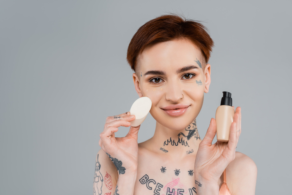 happy tattooed woman holding makeup foundation and sponge isolated on grey - Photo, Image