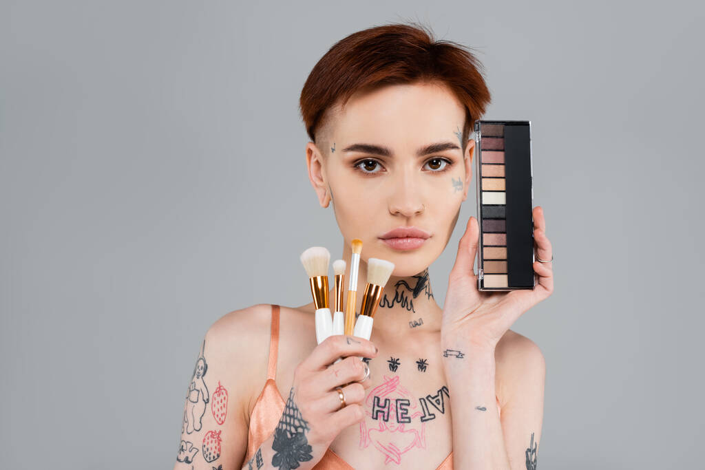 young tattooed woman holding eye shadow palette and cosmetic brushes isolated on grey - Photo, Image