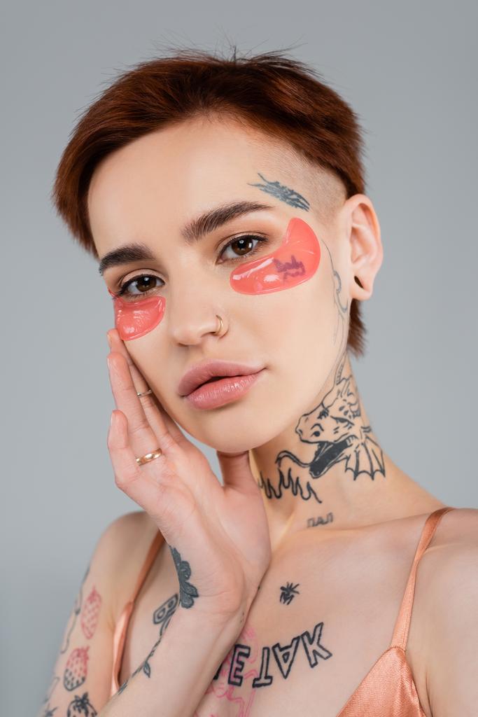 mulher jovem e tatuada com manchas hidratantes nos olhos isoladas em cinza - Foto, Imagem