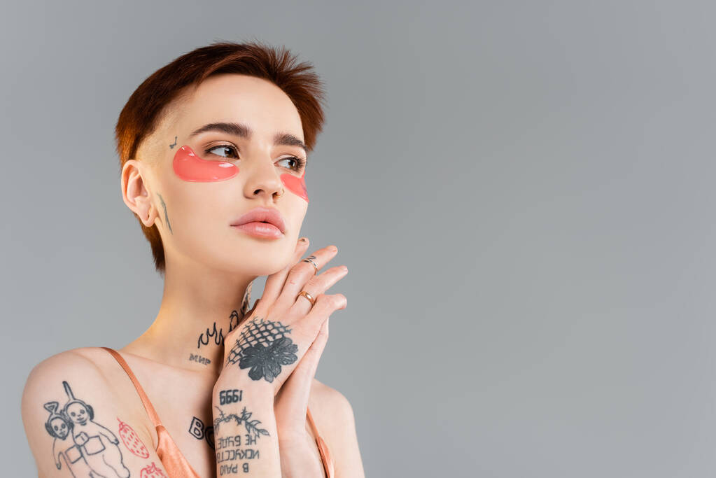 νεαρή γυναίκα με τατουάζ σε ενυδατικά μπαλώματα ματιών κοιτάζοντας μακριά απομονωμένη σε γκρι - Φωτογραφία, εικόνα