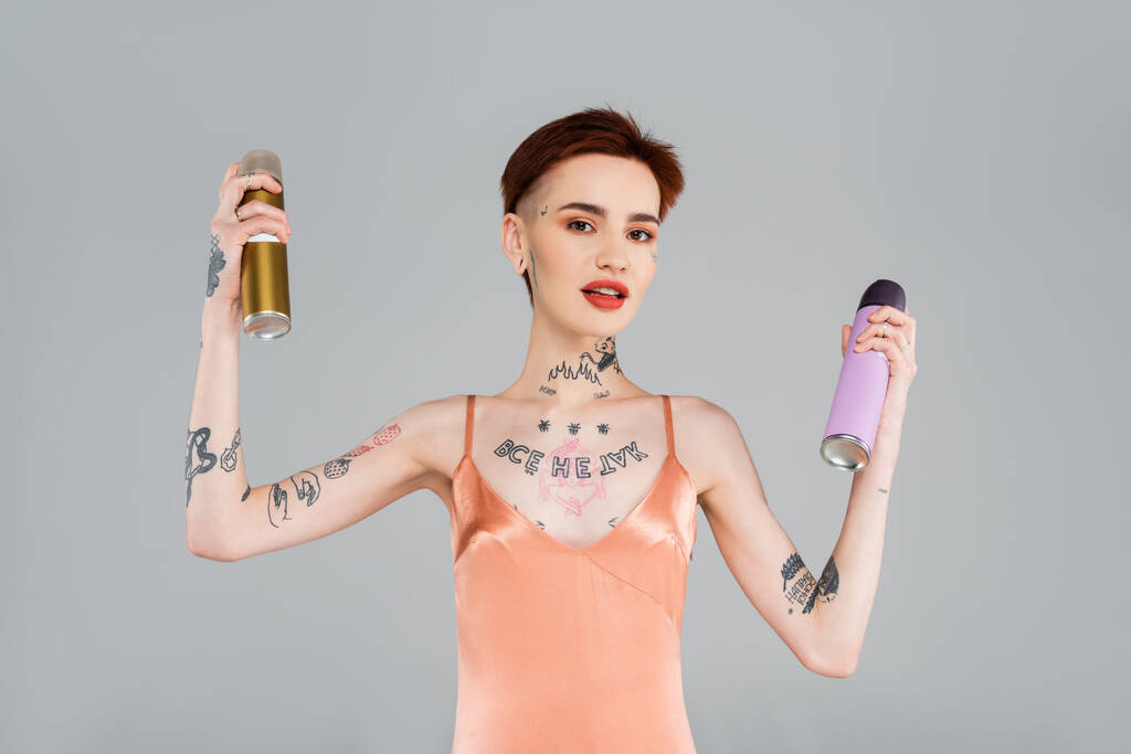 mujer joven tatuada con labios rojos sosteniendo botellas de laca aisladas en gris - Foto, imagen