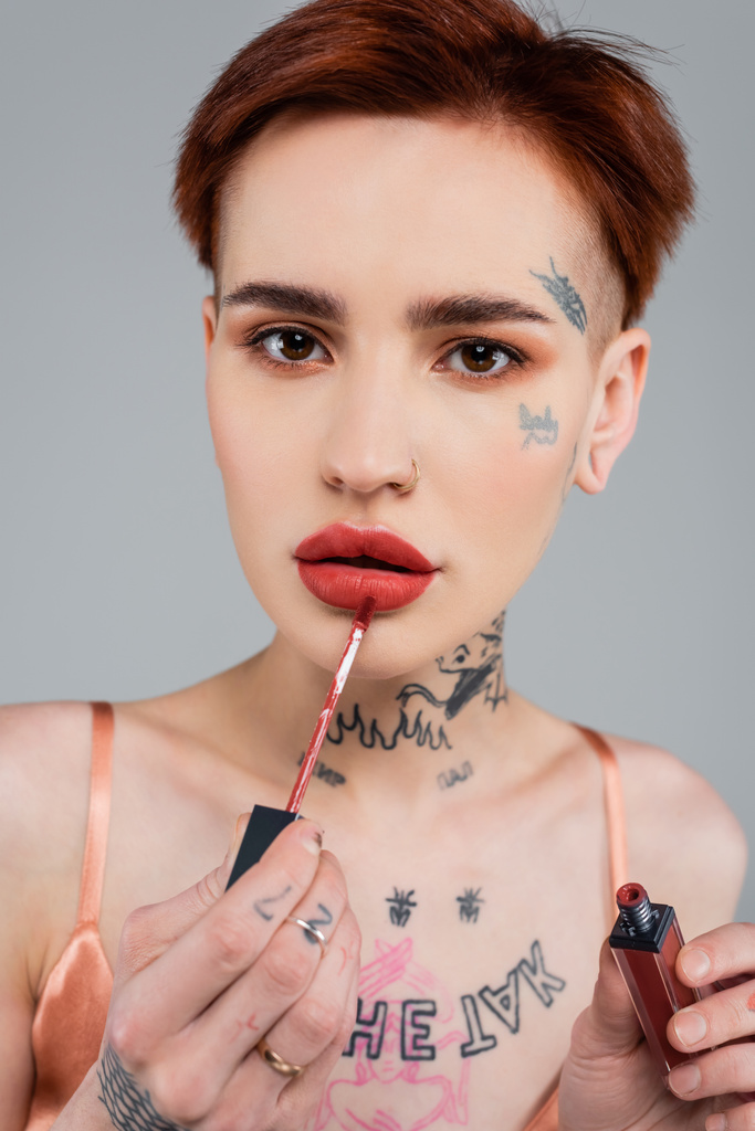 татуированная женщина с красными губами наносит блеск для губ, изолированный на сером - Фото, изображение