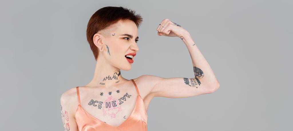 tatuoitu nuori nainen punaiset huulet osoittaa lihas eristetty harmaa, banneri - Valokuva, kuva