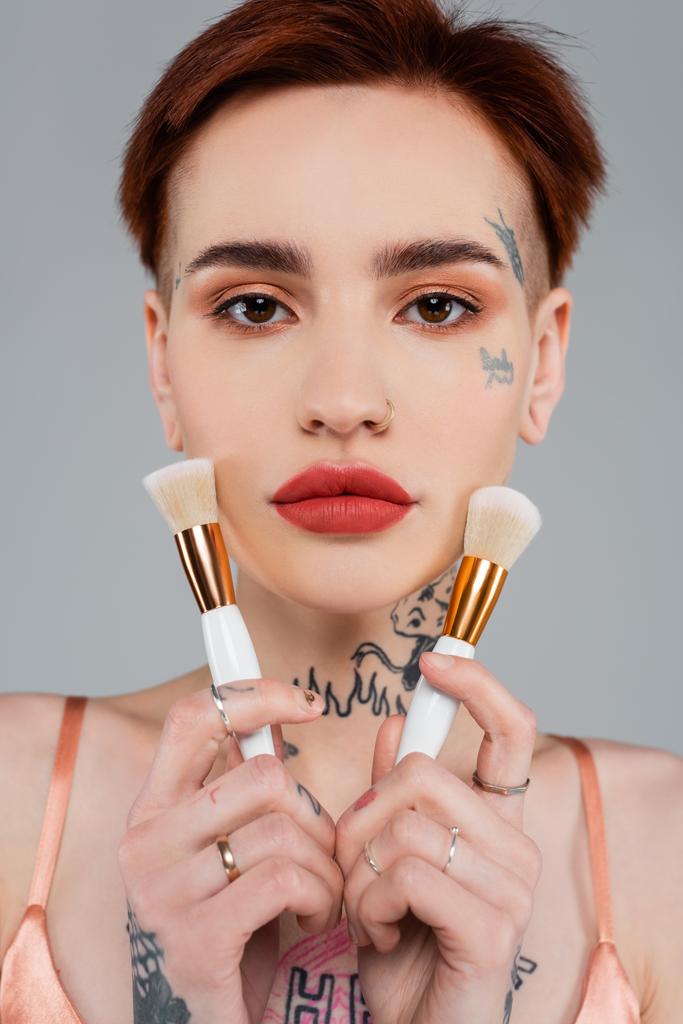 jeune femme tatouée avec des lèvres rouges tenant des pinceaux cosmétiques près du visage isolé sur gris - Photo, image