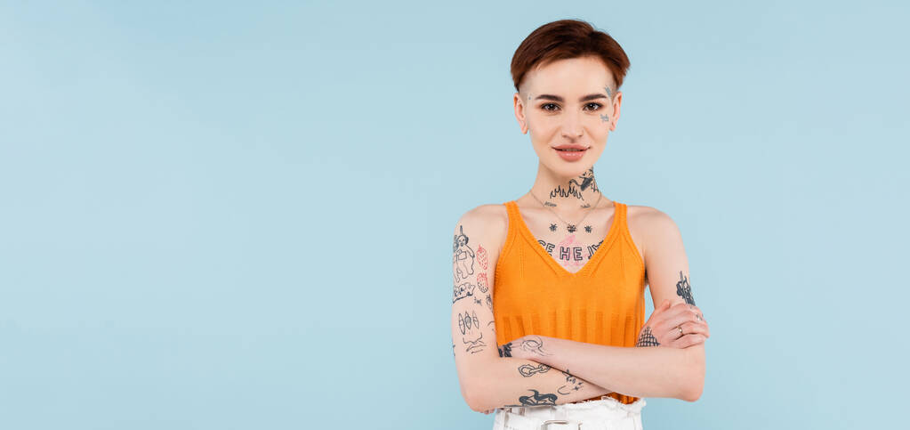 donna soddisfatta e tatuata in maglia arancione canotta in piedi con braccia incrociate isolate su blu, banner - Foto, immagini