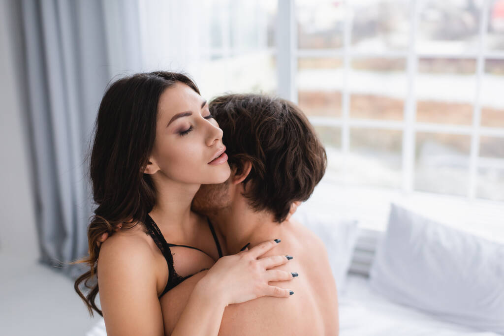 Sensual woman in bra hugging boyfriend in blurred bedroom  - Photo, Image