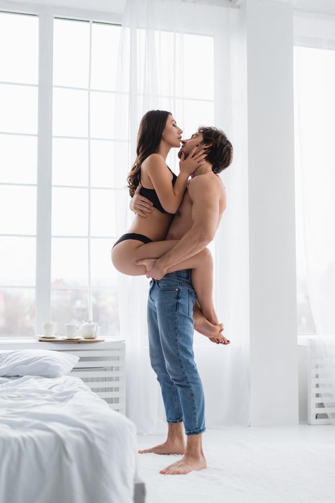 Вид сбоку на мужчину в джинсах, держащего сексуальную девушку в спальне  - Фото, изображение