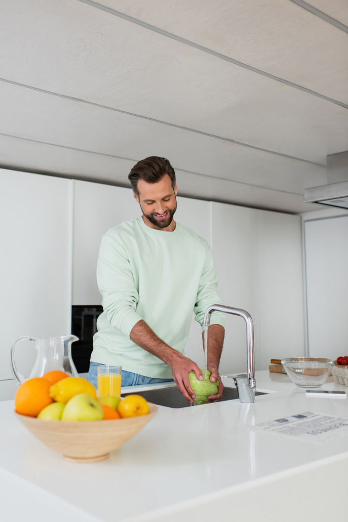 Lächelnder Mann wäscht Salat, während er das Frühstück in der Nähe von frischem Obst zubereitet - Foto, Bild