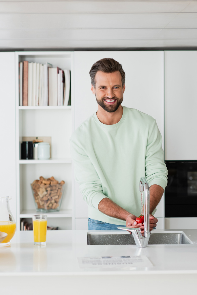 uomo sorridente alla macchina fotografica mentre lavava i pomodorini in cucina - Foto, immagini