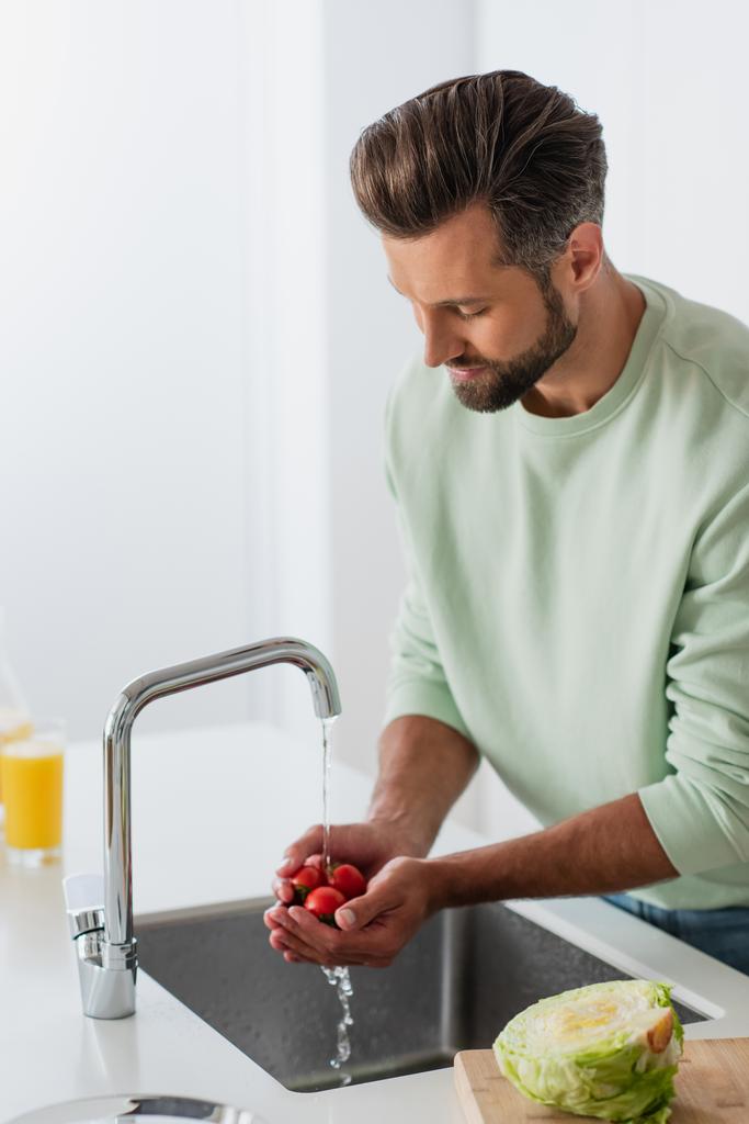 człowiek mycie dojrzałych pomidorów wiśniowych w pobliżu świeżej sałaty w kuchni - Zdjęcie, obraz