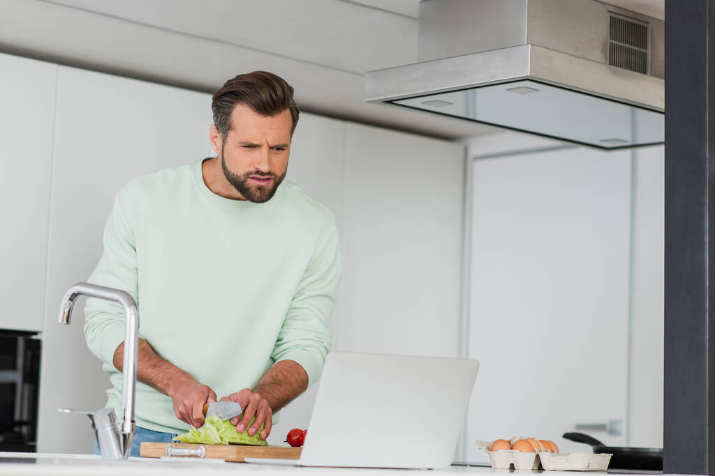 άνθρωπος κοπής λαχανικών, ενώ προετοιμασία πρωινό κοντά στο φορητό υπολογιστή στην κουζίνα - Φωτογραφία, εικόνα
