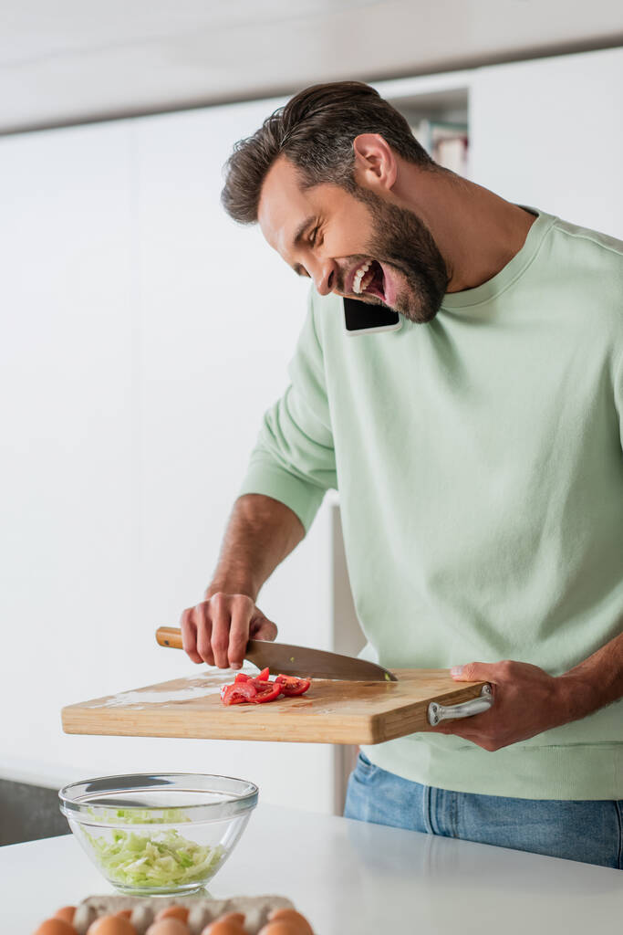 Aufgeregter Mann lacht, während er frischen Salat zubereitet und mit dem Smartphone spricht - Foto, Bild