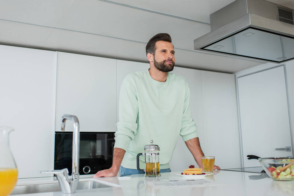 positieve man kijkt weg in de buurt van groene thee en heerlijke pannenkoeken in de keuken - Foto, afbeelding