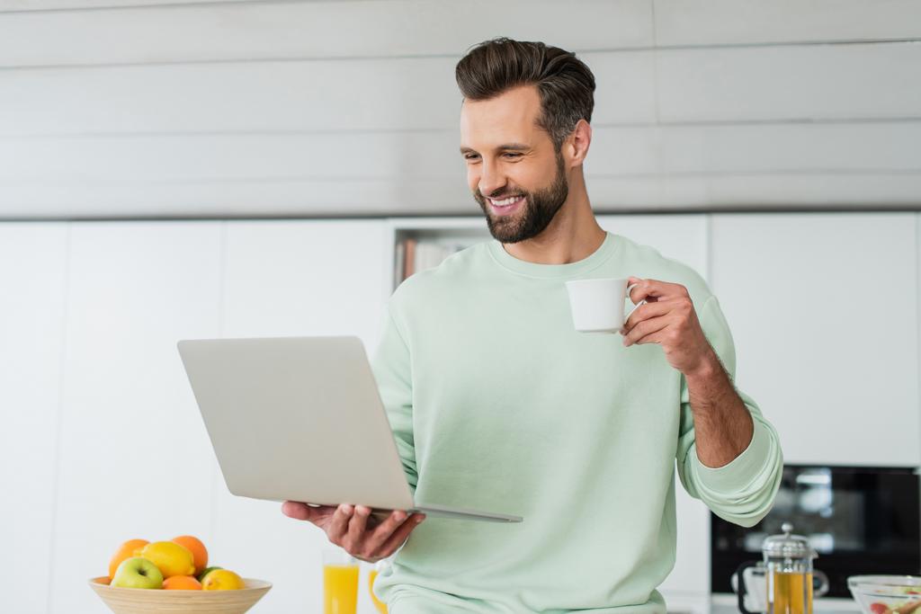 χαρούμενος άνθρωπος κοιτάζοντας φορητό υπολογιστή, ενώ πίνοντας καφέ στην κουζίνα - Φωτογραφία, εικόνα
