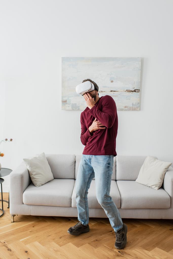 hombre asustado en vr auriculares cubriendo la boca con la mano mientras juega en casa - Foto, Imagen
