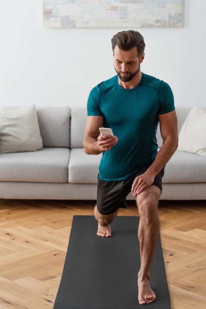 Босоногий мужчина смотрит на смартфон, практикуя йогу дома - Фото, изображение