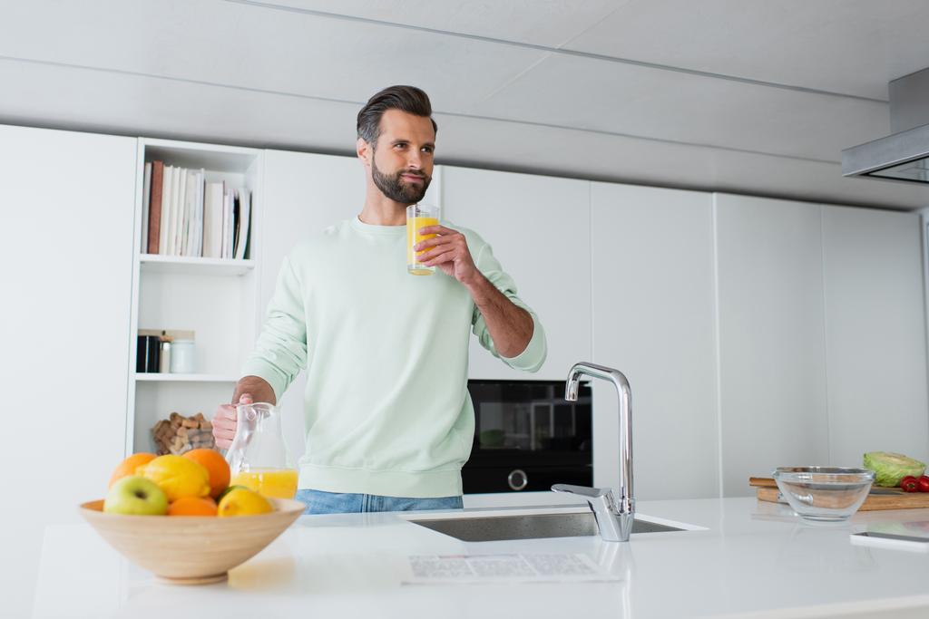 Mutfakta taze meyvelerin yanında portakal suyu içen memnun bir adam. - Fotoğraf, Görsel