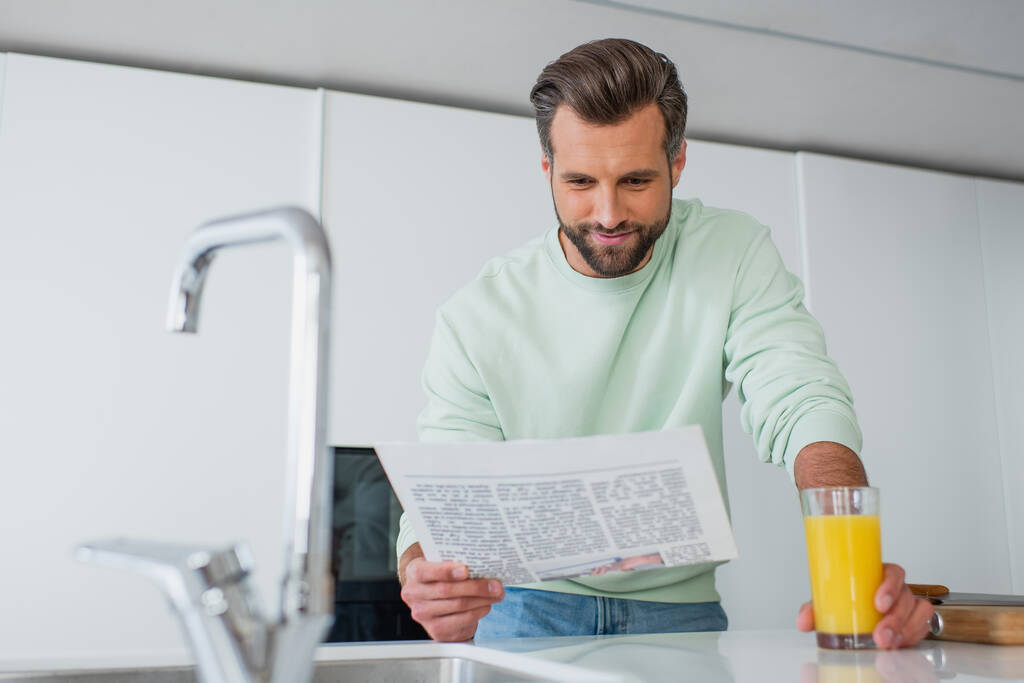 positieve man leest krant in de buurt van glas sinaasappelsap en kraan op wazig voorgrond - Foto, afbeelding
