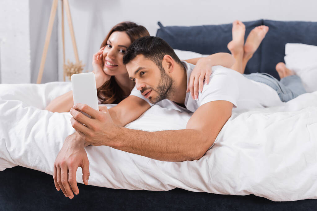 Smartphone in hand of blurred man taking selfie with girlfriend on bed  - Photo, Image